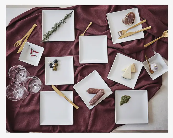 Et sæt firkantede hvide keramiktallerkener er arrangeret på en vinrød dug, hvor hver tallerken viser forskellige madvarer såsom urter, skiveskåret kød, oste, bær og saucer. Tallerkenerne varierer i størrelse og er ledsaget af guldfarvet bestik og klare vinglas. Den samlede præsentation er elegant og velegnet til servering af forretter eller små retter.